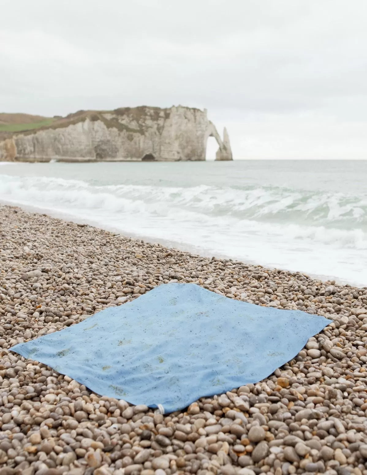 Carré de soie biologique bleu posé sur une plage de galets