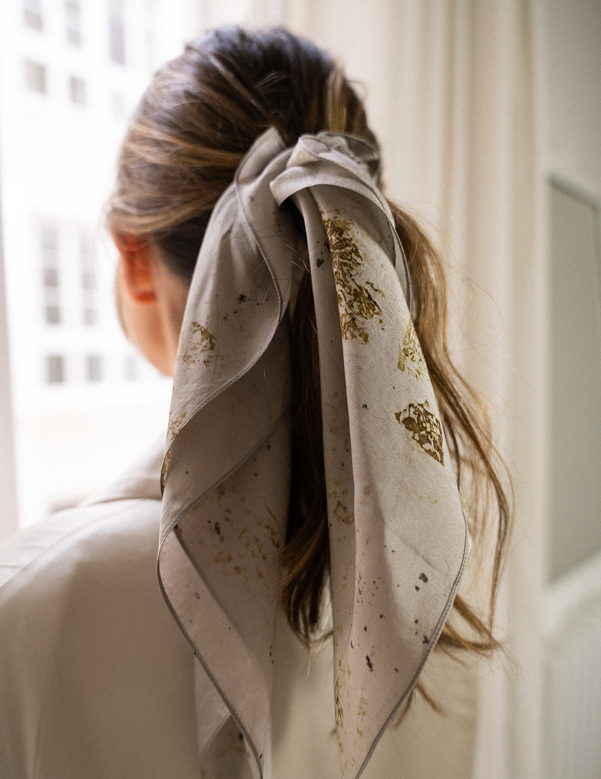 Zoom sur un Chouchou-foulard en soie porté sur chevelure châtain, en queue de cheval, devant une fenêtre 