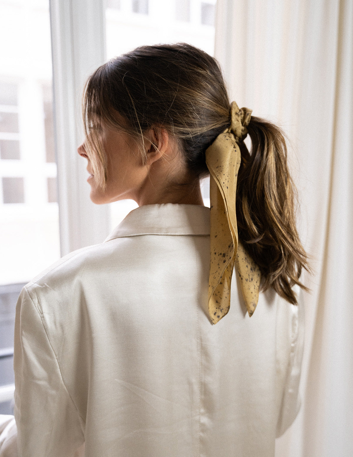 Chouchou-foulard en soie porté sur chevelure châtain, en queue de cheval, devant une fenêtre, modèle Mudra, teinte dorée 