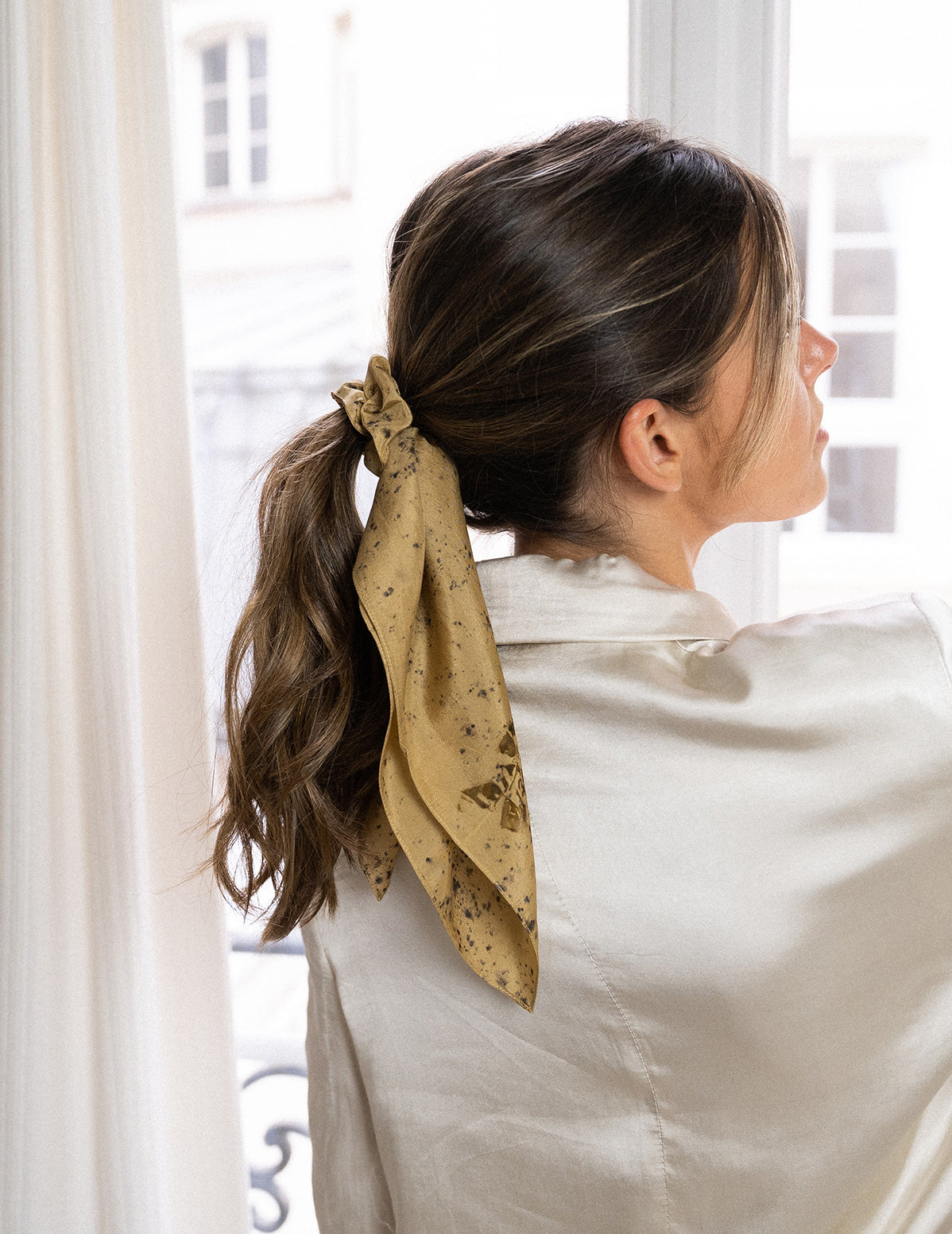 Zoom sur Chouchou-foulard en soie porté sur chevelure châtain, en queue de cheval, devant une fenêtre 